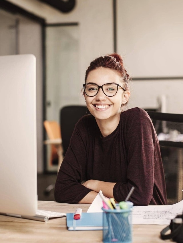Grafikdesigner-Versicherung von Hiscox: Grafikdesignerin sitzt am Schreibtisch und lächelt in die Kamera