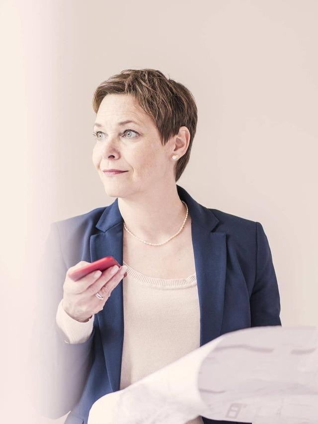 Eine Person in einem blauen Blazer hält ein rotes Smartphone in einer Hand und einige Papiere in der anderen Hand. Das Gesicht der Person ist unscharf, und der Hintergrund ist schlicht und hell, wodurch die Person hervorsticht