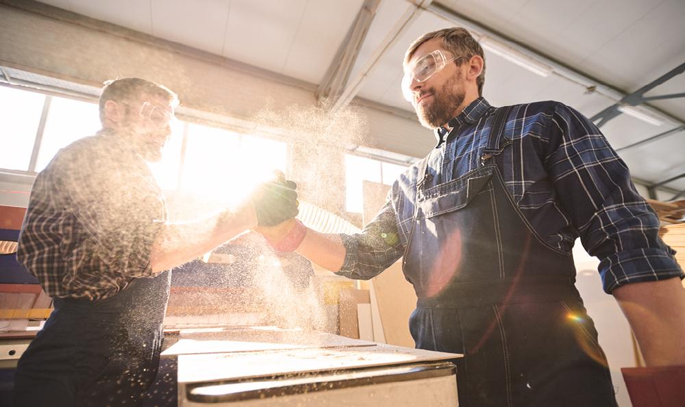 Zwei männliche Handwerker in Schutzkleidung mit Schutzbrillen stehen sich in einer Werkstatt gegenüber und geben sich einen Handschlag. Die Sägespähne fliegen durch die Luft und die Sonne scheint durch das Fenster.