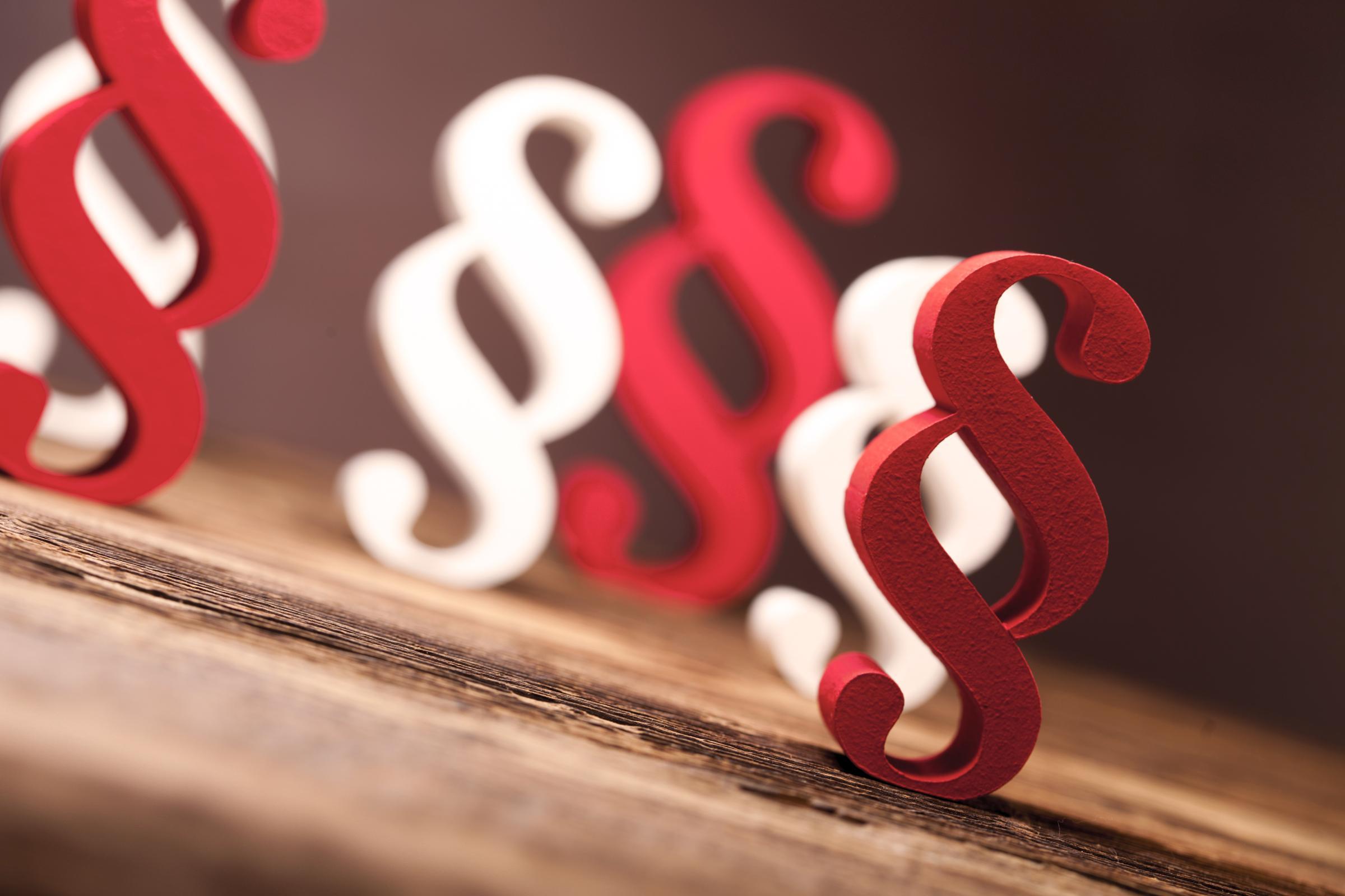 Mehrere rot-weiße Paragrafzeichen in 3D stehen gemischt auf einer Holzfläche.