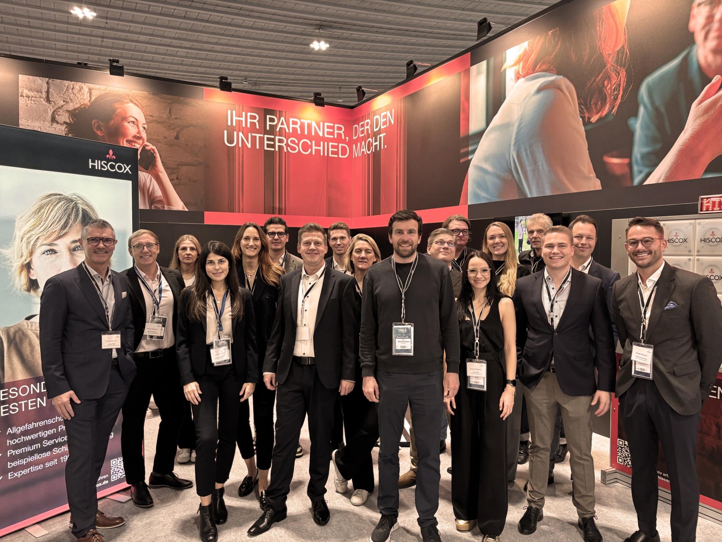 Das große Hiscox Deutschland Team steht in der Gruppe vor dem roten Hiscox Messestand auf der DKM 2024 und lächelt in die Kamera.