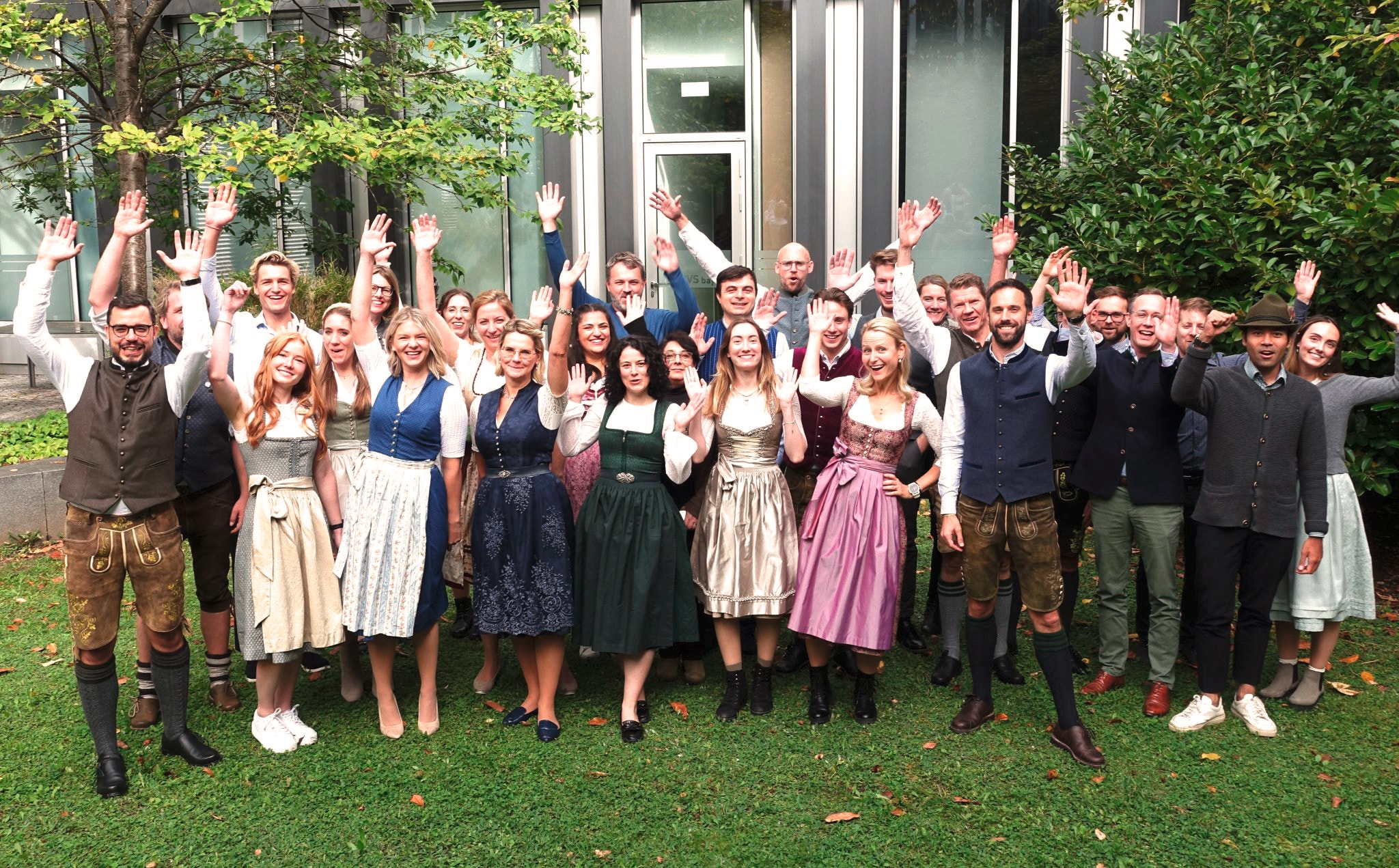 Die Mitarbeiter von Hiscox München stehen in einer großen Gruppe in bayrischer Tracht im Innenhof des Münchner Büros auf der grünen Wiese. Alle haben die Hände und lachen in die Kamera.