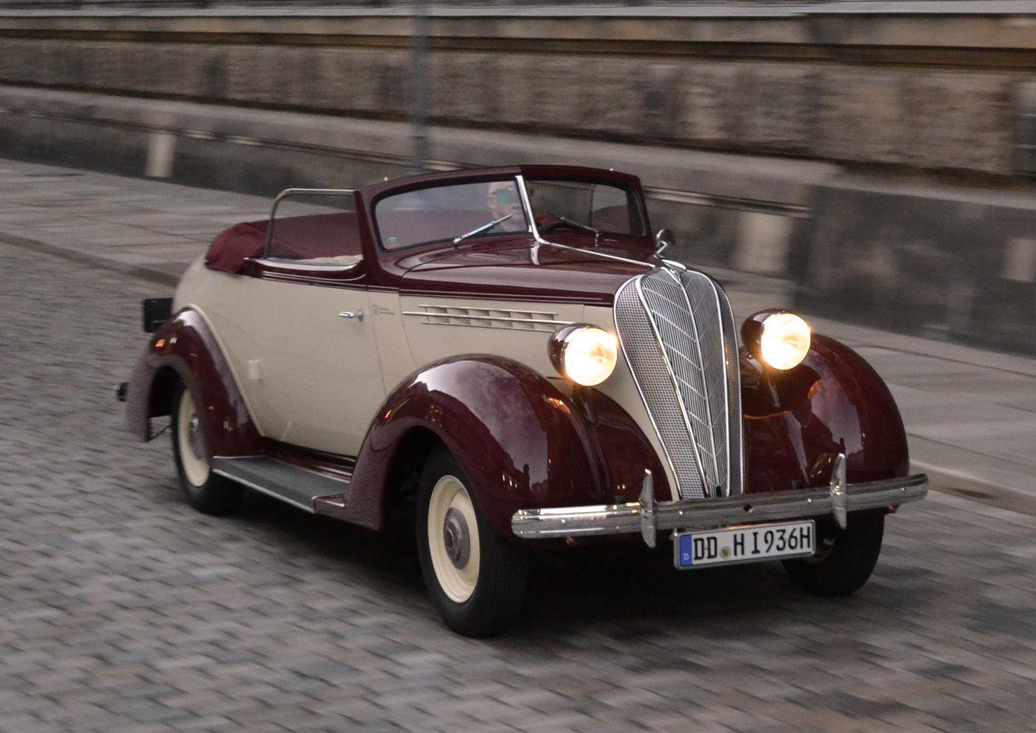 Ein klassisches rot-weißes Cabriolet aus den 1930er Jahren fährt auf einer gepflasterten Straße entlang einer historischen Steinmauer. Das Auto hat runde Scheinwerfer und ein markantes Chromgitter an der Front.