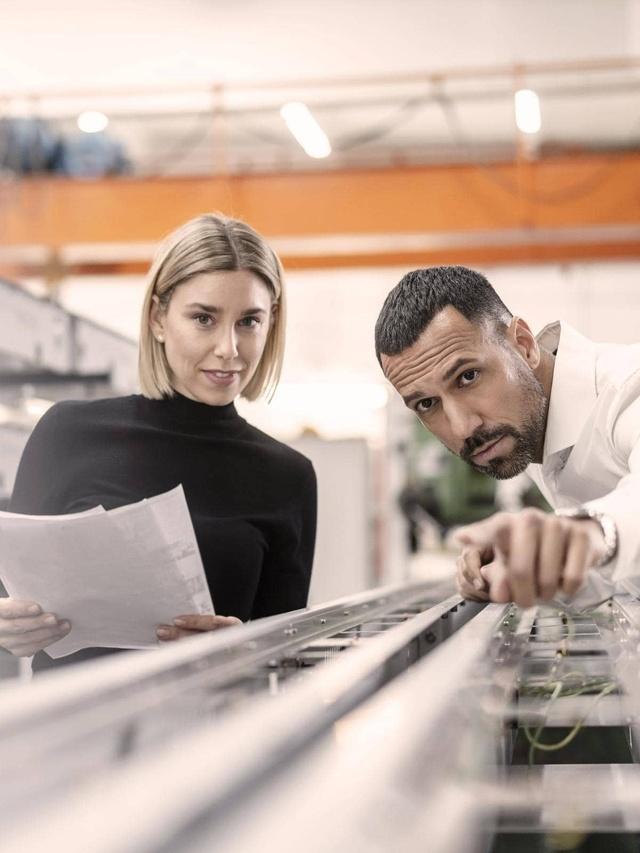 Zwei Personen in einer industriellen Umgebung. Eine Person hält Papiere, die andere zeigt auf eine Maschine. Im Hintergrund sind Fabrikelemente zu sehen.