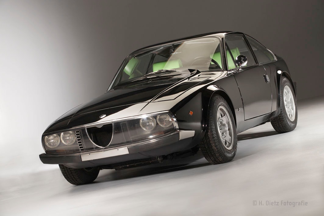 Ein schwarzer Alfa Romeo Montreal von 1970, fotografiert in einem Studio. Das Auto hat runde Scheinwerfer, einen verchromten Kühlergrill und glänzende Karosserie.