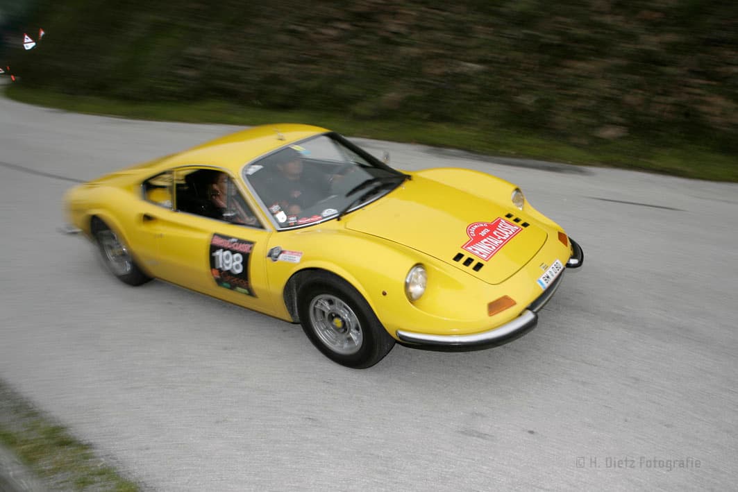 Ein gelbes, klassisches Sportauto mit der Nummer 198, das an einem Rennen teilnimmt. Das Auto fährt auf einer asphaltierten Straße mit grüner Landschaft im Hintergrund.