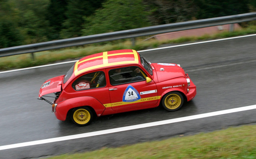Ein rotes Rennfahrzeug, ein Fiat Abarth 1000 TC, fährt auf einer nassen Straße. Das Auto hat die Startnummer 34 und gelbe Felgen. Auf der Motorhaube und dem Dach sind gelbe Streifen zu sehen.