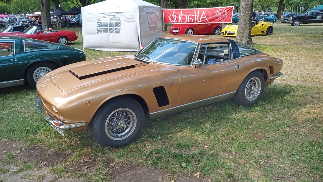 Ein klassisches Auto, ein Iso Grifo, geparkt auf einer Wiese bei einer Autoshow. Das Auto hat eine bronzene Farbe mit schwarzen Akzenten und Speichenrädern.