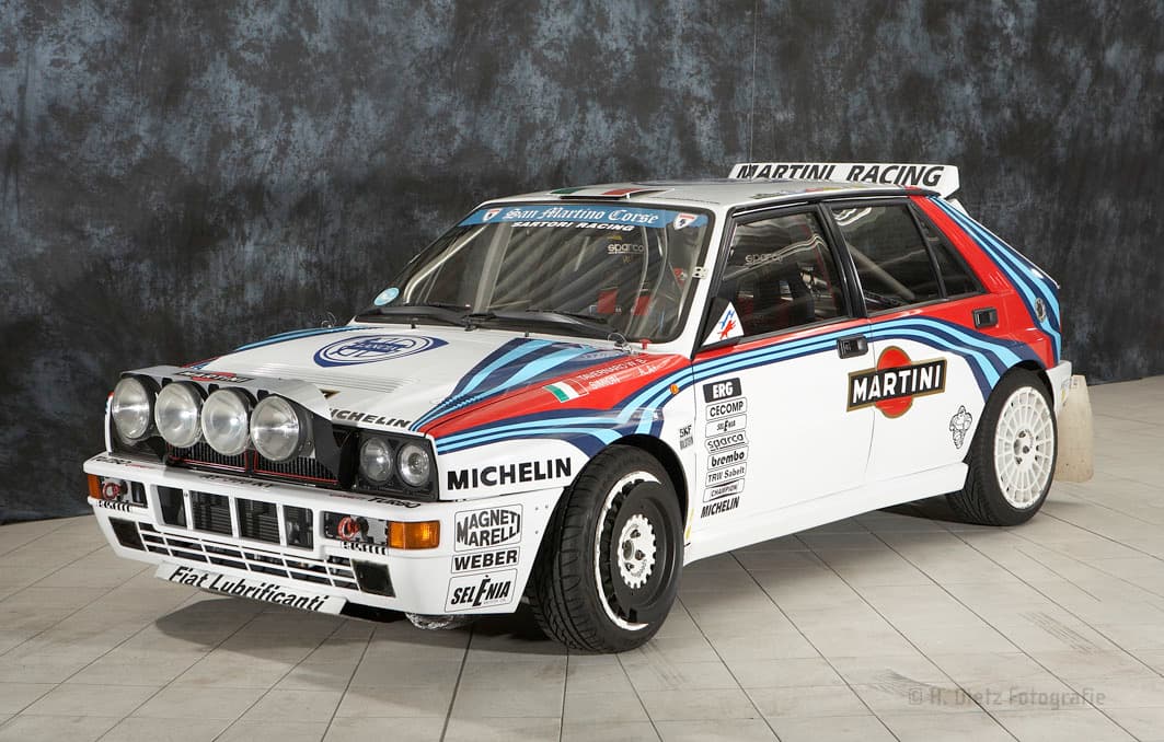 Ein weißer Rallye-Wagen mit blauen und roten Streifen, gesponsert von Martini Racing. Das Auto hat mehrere Aufkleber von Sponsoren wie Michelin und Magneti Marelli. Es ist auf einem grauen Fliesenboden in einem Innenraum geparkt.