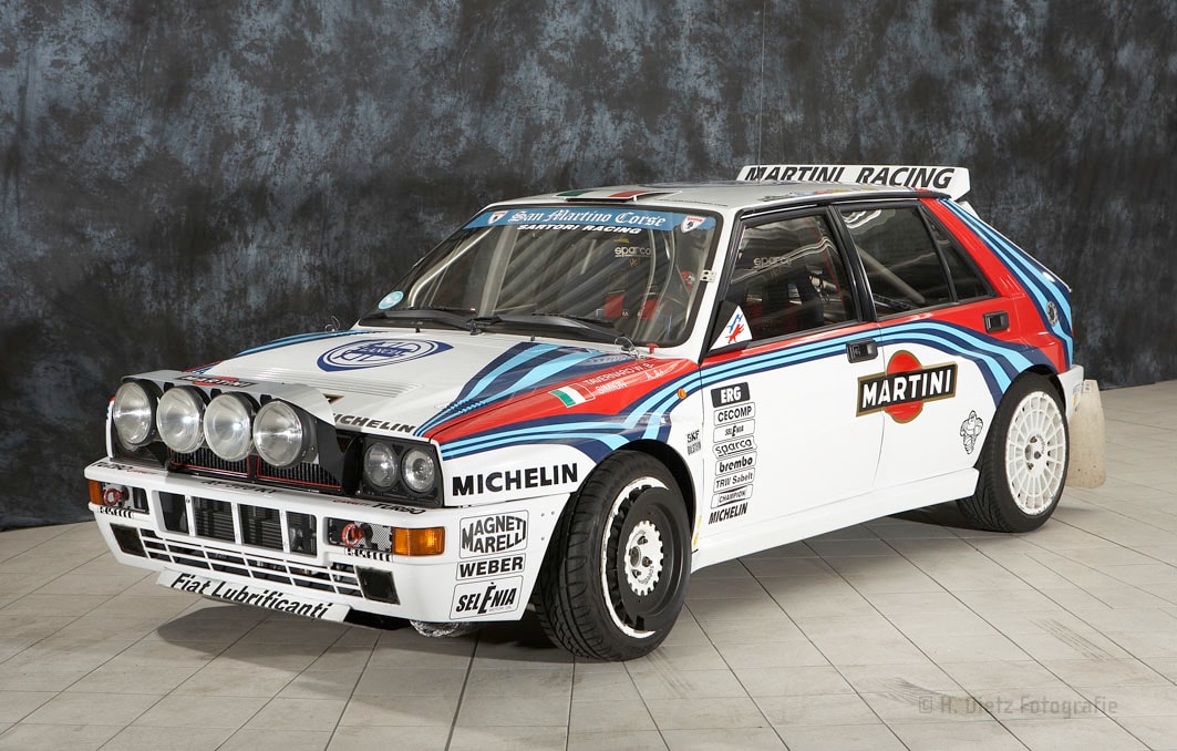 Ein weißer Rallye-Wagen mit blauen und roten Streifen, gesponsert von Martini Racing. Das Auto hat mehrere Aufkleber von Sponsoren wie Michelin und Magneti Marelli. Es ist auf einem grauen Fliesenboden in einem Innenraum geparkt.