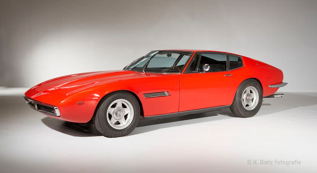 Ein klassischer roter Sportwagen mit aerodynamischem Design, fotografiert in einem Studio. Das Auto hat eine lange Motorhaube, zwei Türen und silberne Räder mit fünf Speichen.