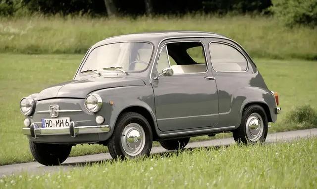 Ein grauer, klassischer Fiat 600, geparkt auf einer Wiese mit Bäumen im Hintergrund. Das Auto hat runde Scheinwerfer, Chromstoßstangen und kleine Räder.