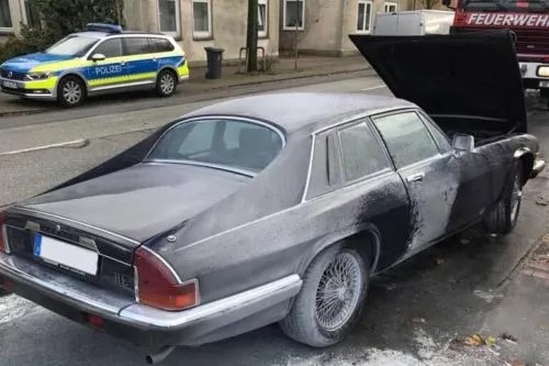 Ein graues Auto mit geöffneter Motorhaube und sichtbaren Schäden am hinteren Teil steht am Straßenrand.