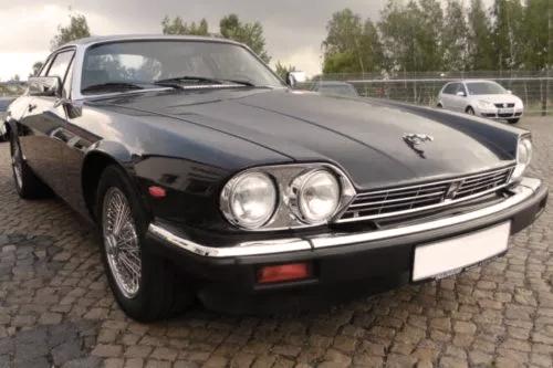 Ein klassischer schwarzer Jaguar XJ-S, geparkt auf einer Kopfsteinpflasterfläche. Das Auto hat markante runde Scheinwerfer, ein schlankes und aerodynamisches Design sowie Chromakzente am Kühlergrill und Stoßfänger.