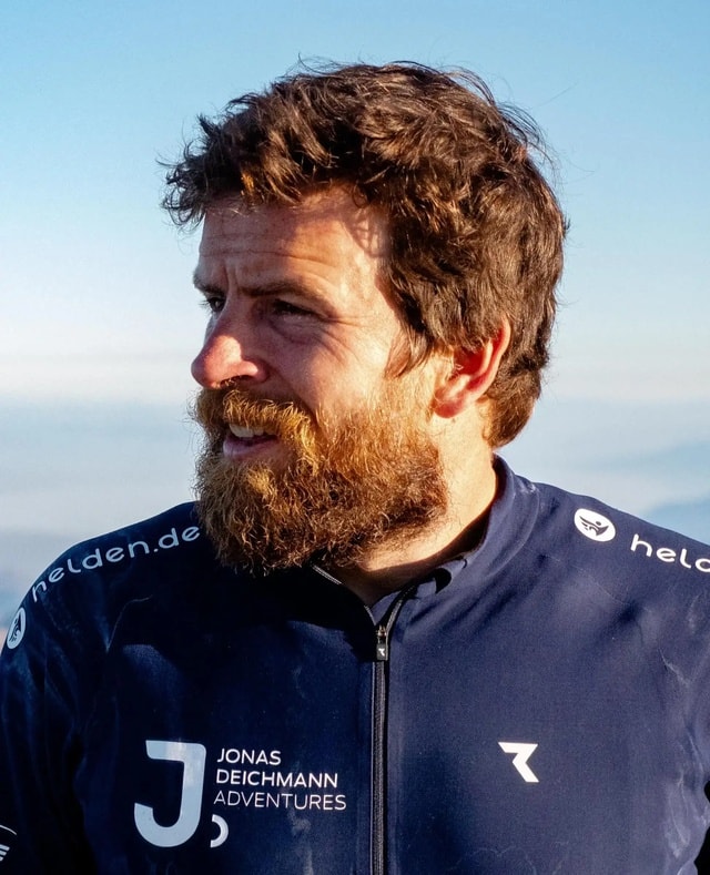 Jonas Deichmann trägt eine dunkelblaue Jacke mit den Aufschriften "Jonas Deichmann Adventures" und "helden.de"