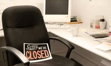 Ein leeres Büro mit einem schwarzen Bürostuhl, auf dem ein Schild mit der Aufschrift "Sorry, WE'RE CLOSED" liegt. Im Hintergrund steht ein alter Computerbildschirm auf einem weißen Schreibtisch, umgeben von verschiedenen Büroartikeln wie Stiften und Papier.