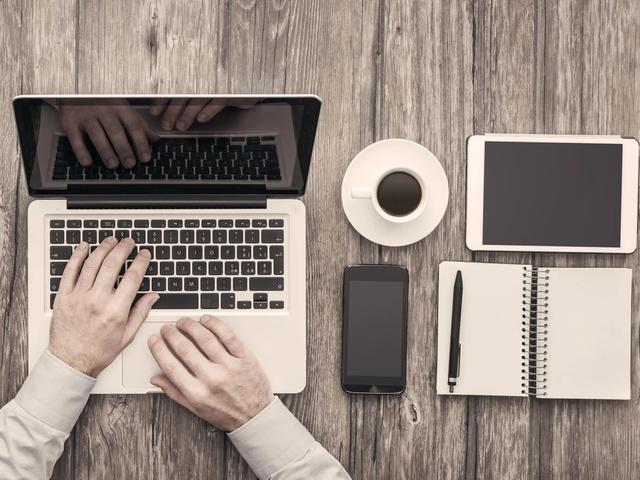 Die Hände einer Person tippen auf einem Laptop. Rechts auf dem Tisch sind eine Kaffeetasse, ein Tablet, ein Smartphone und ein Notizbuch mit einem Stift.