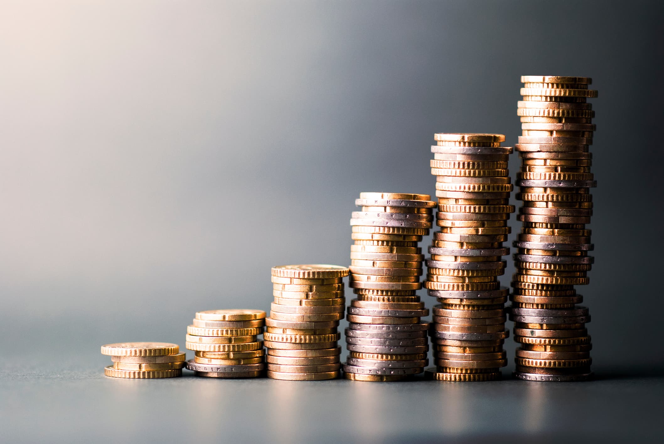 Gestapelte Münzen in verschiedenen Höhen, die von links nach rechts ansteigen. Die Münztürme symbolisieren ein wachsendes Vermögen. Hintergrund ist schlicht und grau gehalten.
