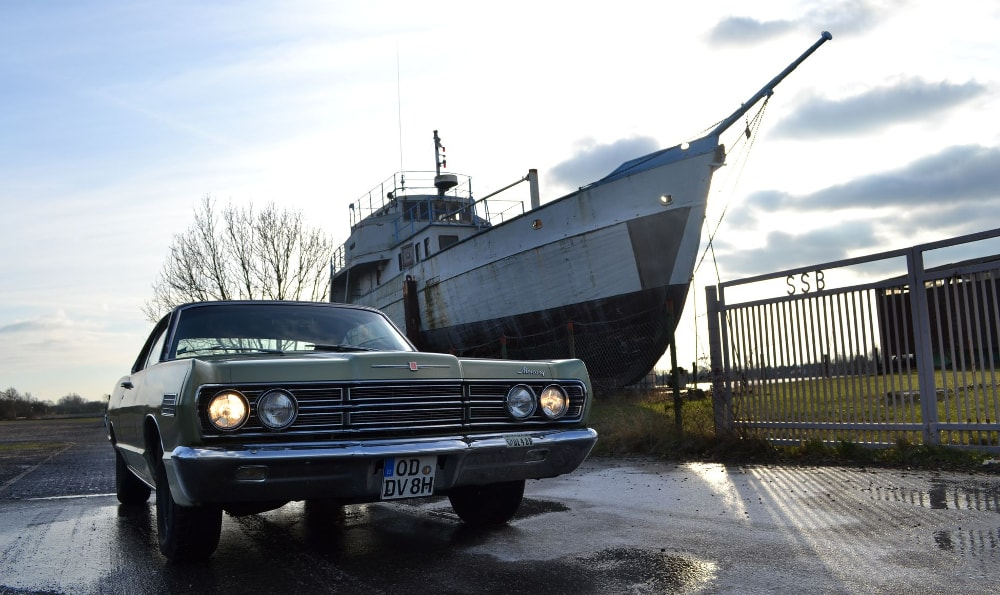 Ein klassisches Auto steht auf einer nassen Straße, mit einem großen, alten Schiff im Hintergrund. Das Auto hat das Nummernschild "OD DV 8H" und die Szene spielt vermutlich am späten Nachmittag oder frühen Abend.