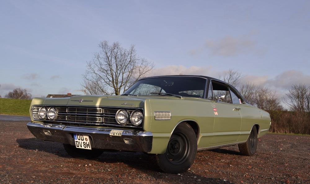 Ein klassischer grüner Ford Galaxie 500 ist auf einer Kiesfläche geparkt. Der Himmel ist klar und im Hintergrund sind einige Bäume zu sehen. Das Auto hat Chromdetails, runde Scheinwerfer und ein schwarzes Dach.