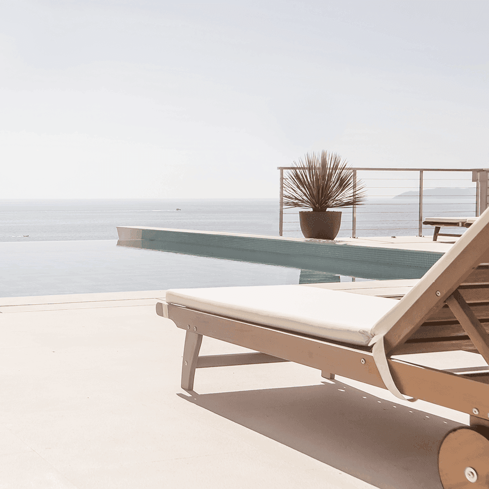 Sonnenliege aus Holz steht an einem Infinity-Pool mit Pflanze und Meer im Hintergrund.