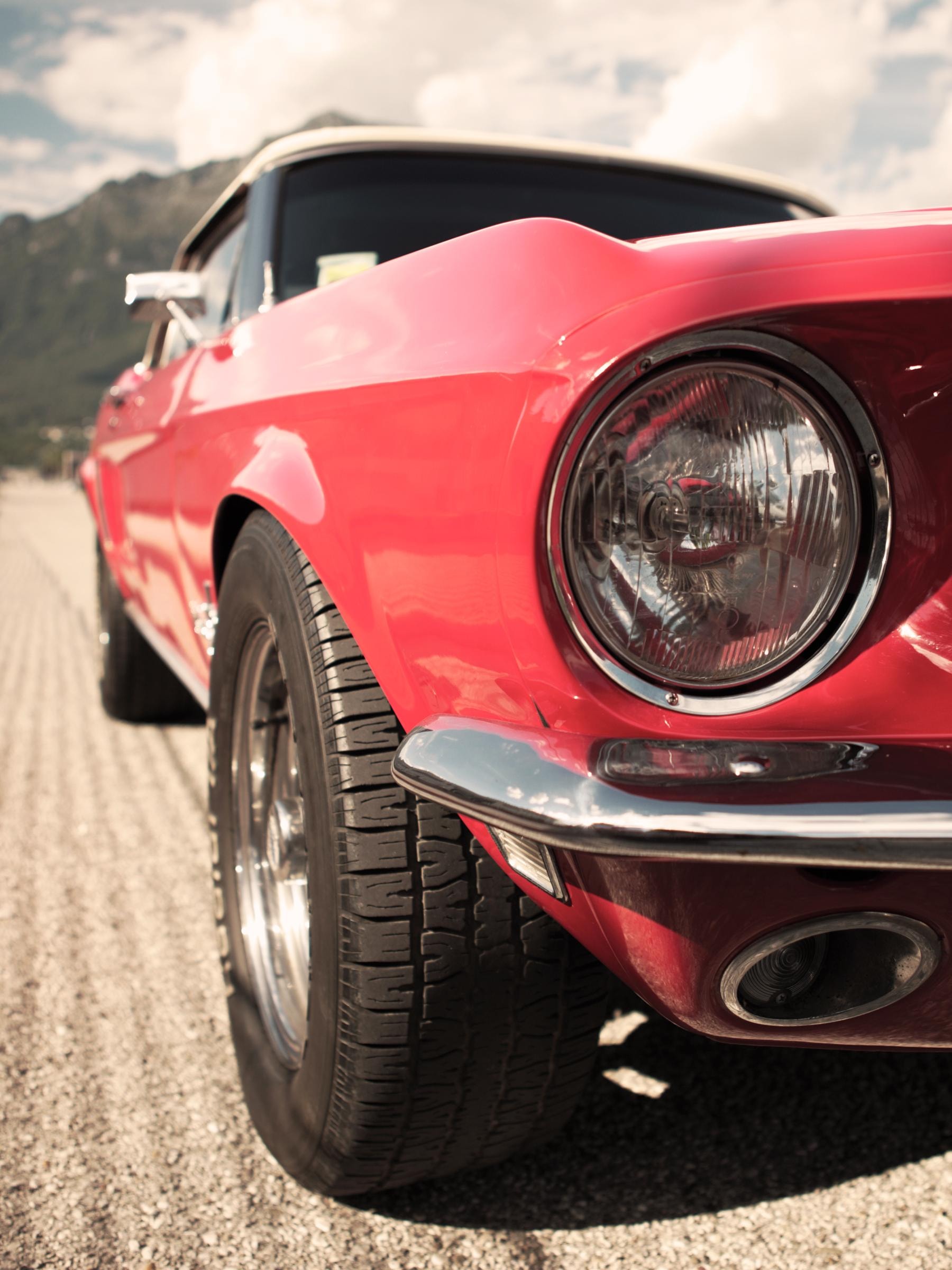 Nahaufnahme der Vorderseite eines klassischen roten Autos, wahrscheinlich ein Vintage-Modell. Der Fokus liegt auf dem Scheinwerfer, dem Reifen und einem Teil des Kotflügels. Im Hintergrund ist eine bergige Landschaft unter einem teilweise bewölkten Himmel zu sehen.