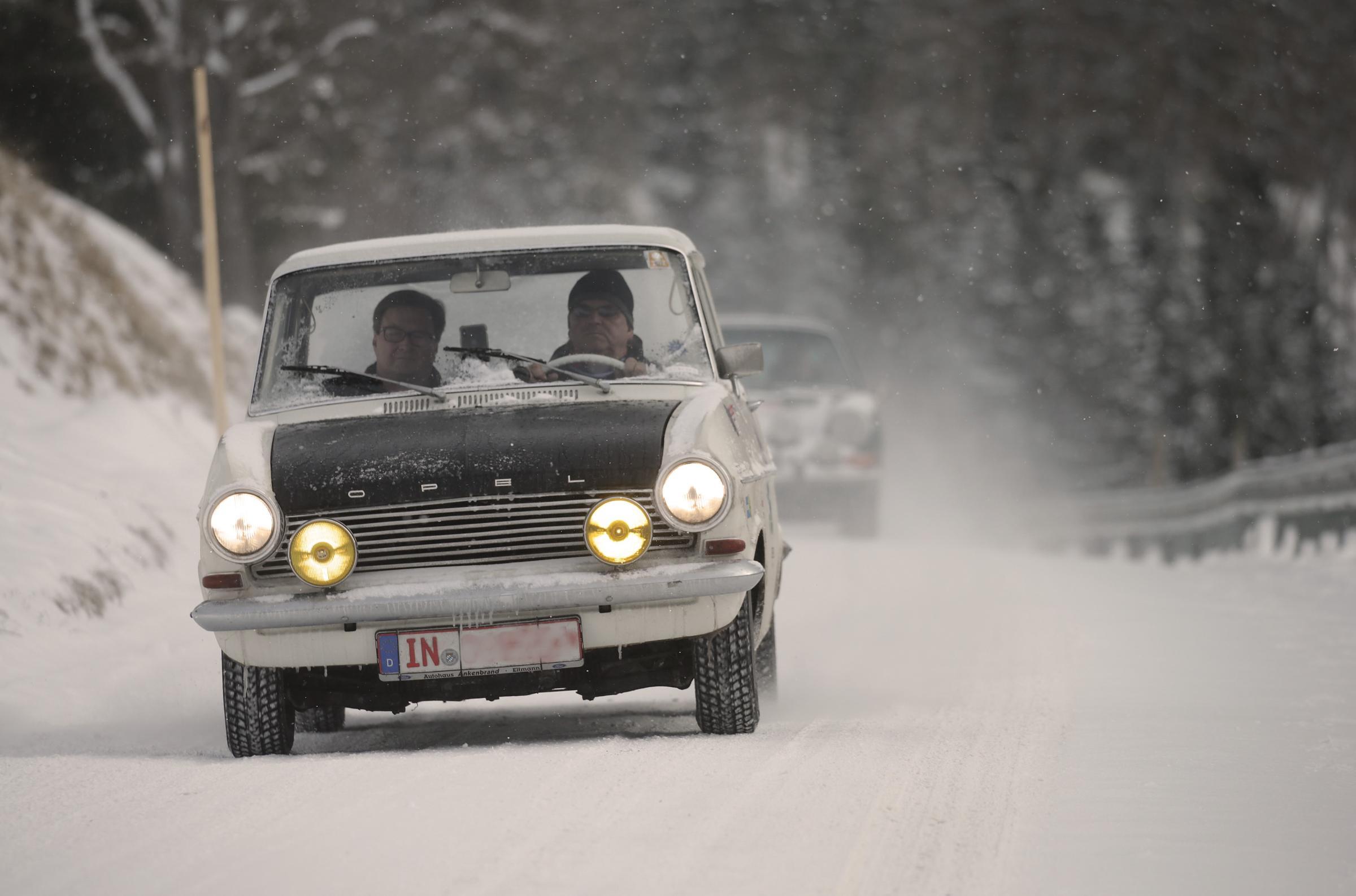 Ein klassisches Ford-Auto fährt auf einer schneebedeckten Straße, gefolgt von einem weiteren Fahrzeug. Die Szene spielt in einem verschneiten Waldgebiet, was auf Winterbedingungen hinweist, und die Scheinwerfer des Autos sind eingeschaltet, und das Nummernschild zeigt teilweise "IN"