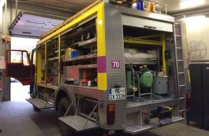 Ein gelbes Feuerwehrfahrzeug steht in einer Garage. Die hintere Tür des Fahrzeugs ist geöffnet und zeigt verschiedene Ausrüstungsgegenstände, darunter Schläuche, Werkzeuge und ein grüner Generator.