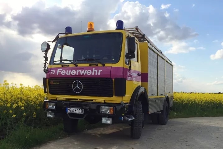 Ein gelbes Feuerwehrauto mit der Aufschrift "Feuerwehr" auf einem roten Streifen an der Front. Es hat ein Mercedes-Benz-Logo und blaue sowie orangefarbene Lichter auf dem Dach. Das Fahrzeug steht auf einem Feldweg neben einem Feld mit gelben Blumen.