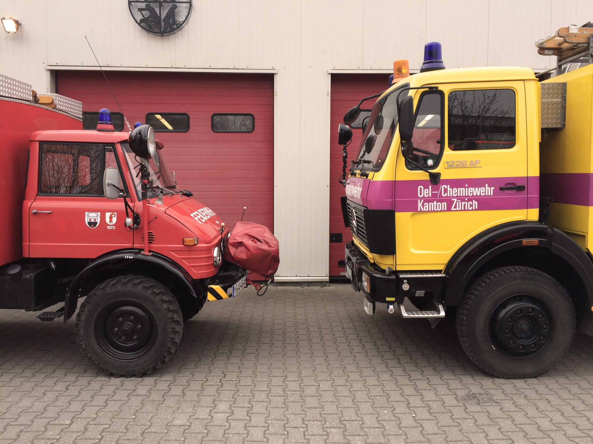 Ein rotes Feuerwehrauto und ein gelbes Feuerwehrauto stehen sich vor einem Gebäude mit roten Garagentoren gegenüber.