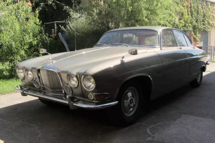 Ein klassischer Jaguar Mark X mit langer Motorhaube, Chromakzenten und runden Scheinwerfern, geparkt auf einer Auffahrt mit Grünfläche im Hintergrund.