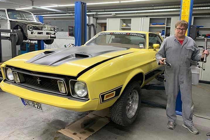 Ein gelber klassischer Ford Mustang in einer Werkstatt, der auf einer Hebebühne steht, und eine Person steht daneben und hält Werkzeuge.