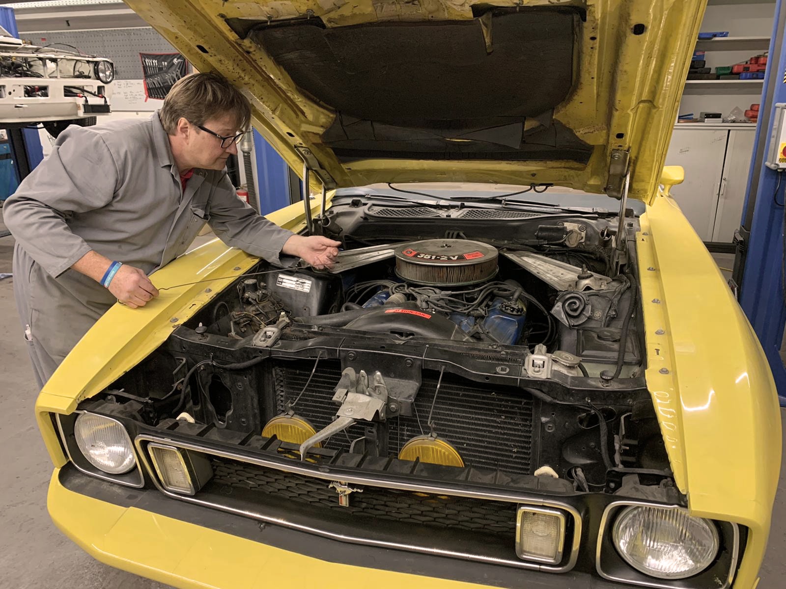 Ein Mechaniker in grauer Arbeitskleidung arbeitet an einem gelben Oldtimer mit geöffneter Motorhaube.