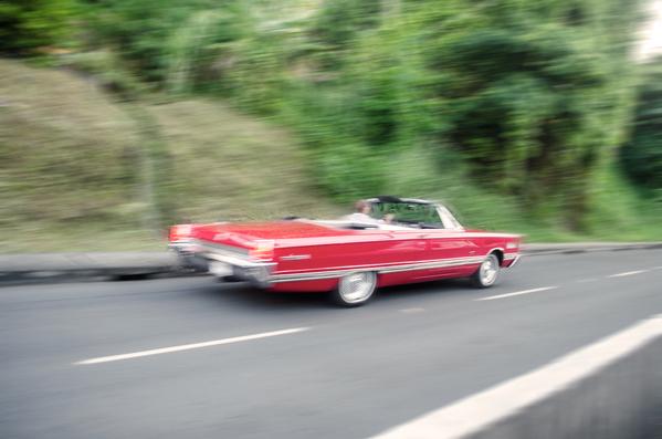 Ein rotes Cabrio fährt auf einer Straße mit verschwommenem Grün im Hintergrund, was auf Bewegung hinweist.