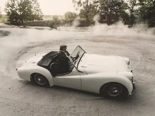 Ein weißes Cabrio driftet auf einer Schotterfläche und wirbelt Staub auf. Im Hintergrund sind Bäume und Grünflächen zu sehen.