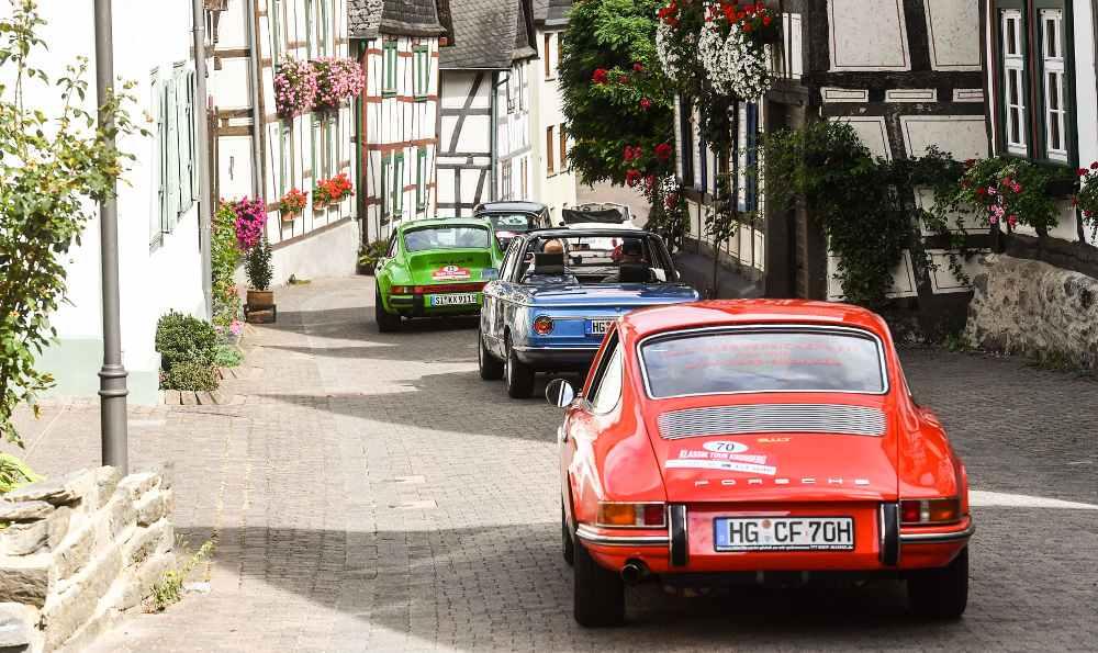 Eine Reihe von Oldtimern fährt durch eine enge Straße in einem europäischen Dorf mit Fachwerkhäusern und Blumenkästen.