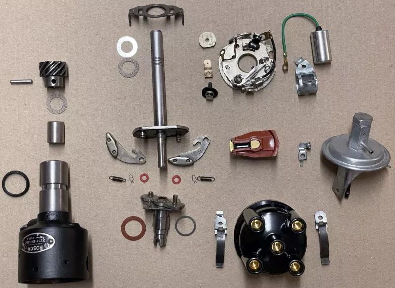 Zerlegter Verteiler eines Verbrennungsmotors mit allen Einzelteilen, wie Verteilerwelle, Rotor, Kappe, Kontakte, Kondensator, sowie mehreren Unterlegscheiben und Schrauben, ordentlich auf einer flachen Oberfläche angeordnet.