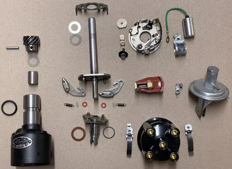 Zerlegter Verteiler eines Verbrennungsmotors mit allen Einzelteilen, wie Verteilerwelle, Rotor, Kappe, Kontakte, Kondensator, sowie mehreren Unterlegscheiben und Schrauben, ordentlich auf einer flachen Oberfläche angeordnet.
