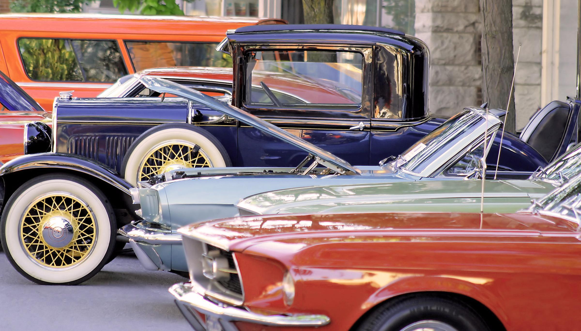 Eine Reihe von Oldtimern, darunter ein orange-rotes Cabrio, ein grünes Auto mit offenem Motor und ein blaues Auto mit Weißwandreifen.