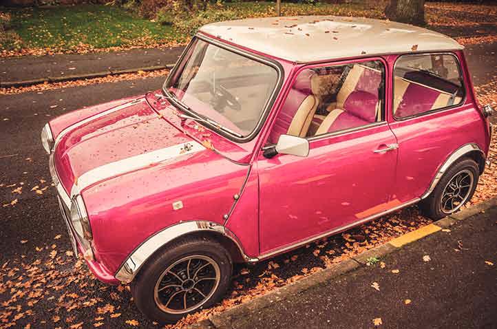 Ein pinkfarbener Mini Cooper mit weißen Streifen steht am Straßenrand, umgeben von herbstlichen Blättern.