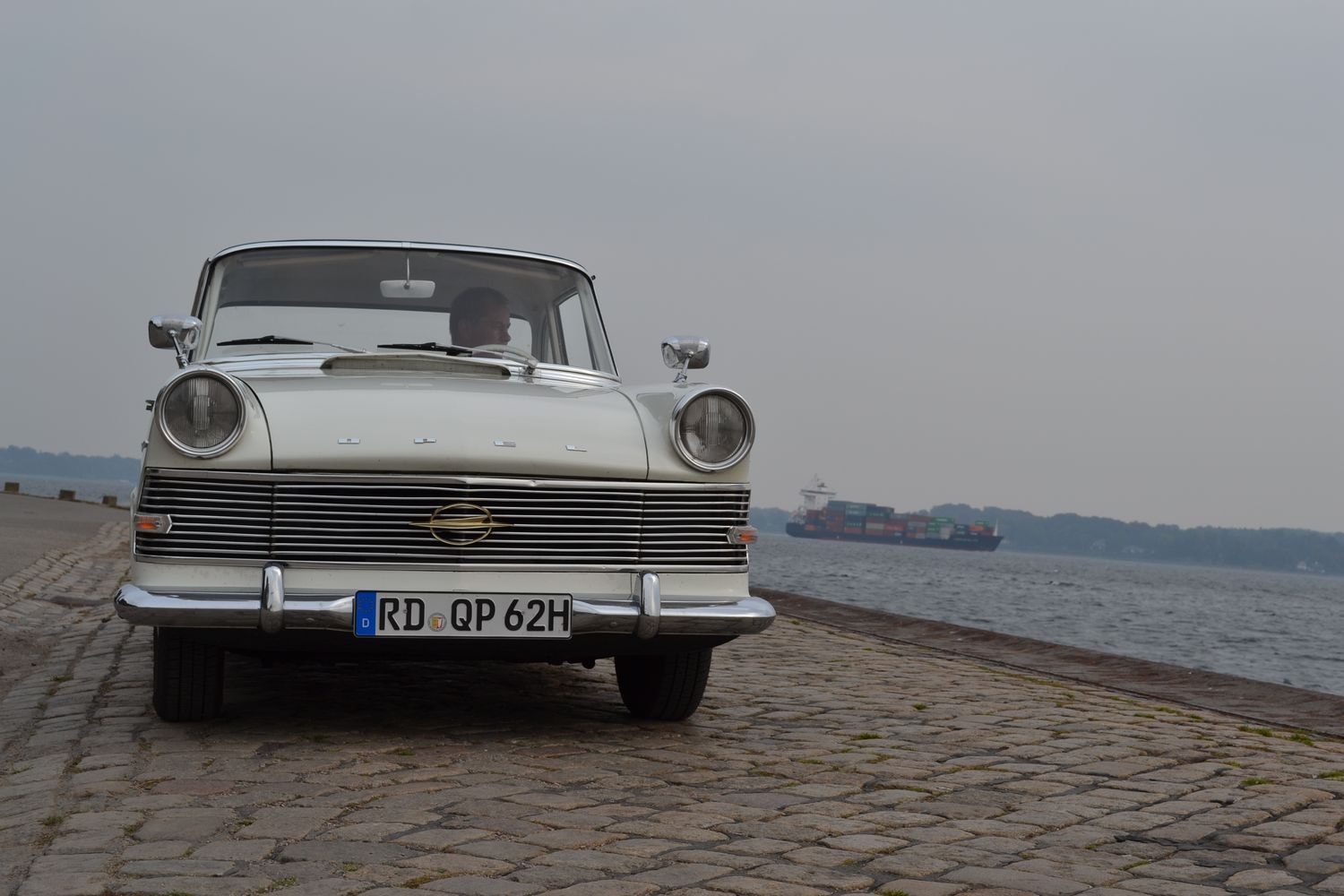Weißes klassisches Auto mit dem Kennzeichen "RD QP 62H" auf einer Kopfsteinpflasterstraße nahe einem Gewässer, im Hintergrund ein großes Frachtschiff.