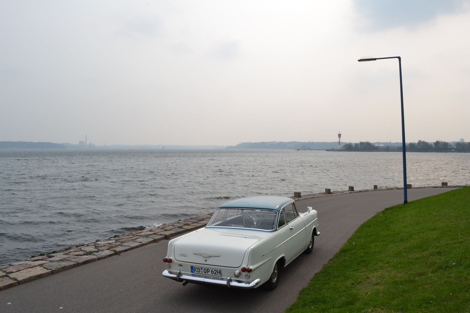Weißes Vintage-Auto mit hellblauem Dach, geparkt auf einer Straße neben einem Gewässer, bei bewölktem Himmel.