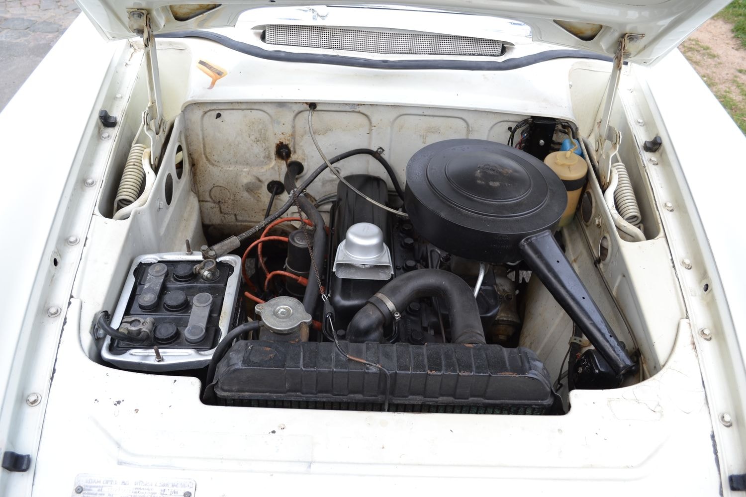 Ein geöffneter Motorraum eines weißen Autos. Der Motor ist sichtbar, mit verschiedenen Kabeln und Komponenten, einschließlich einer Batterie auf der linken Seite und einem Luftfiltergehäuse auf der rechten Seite.