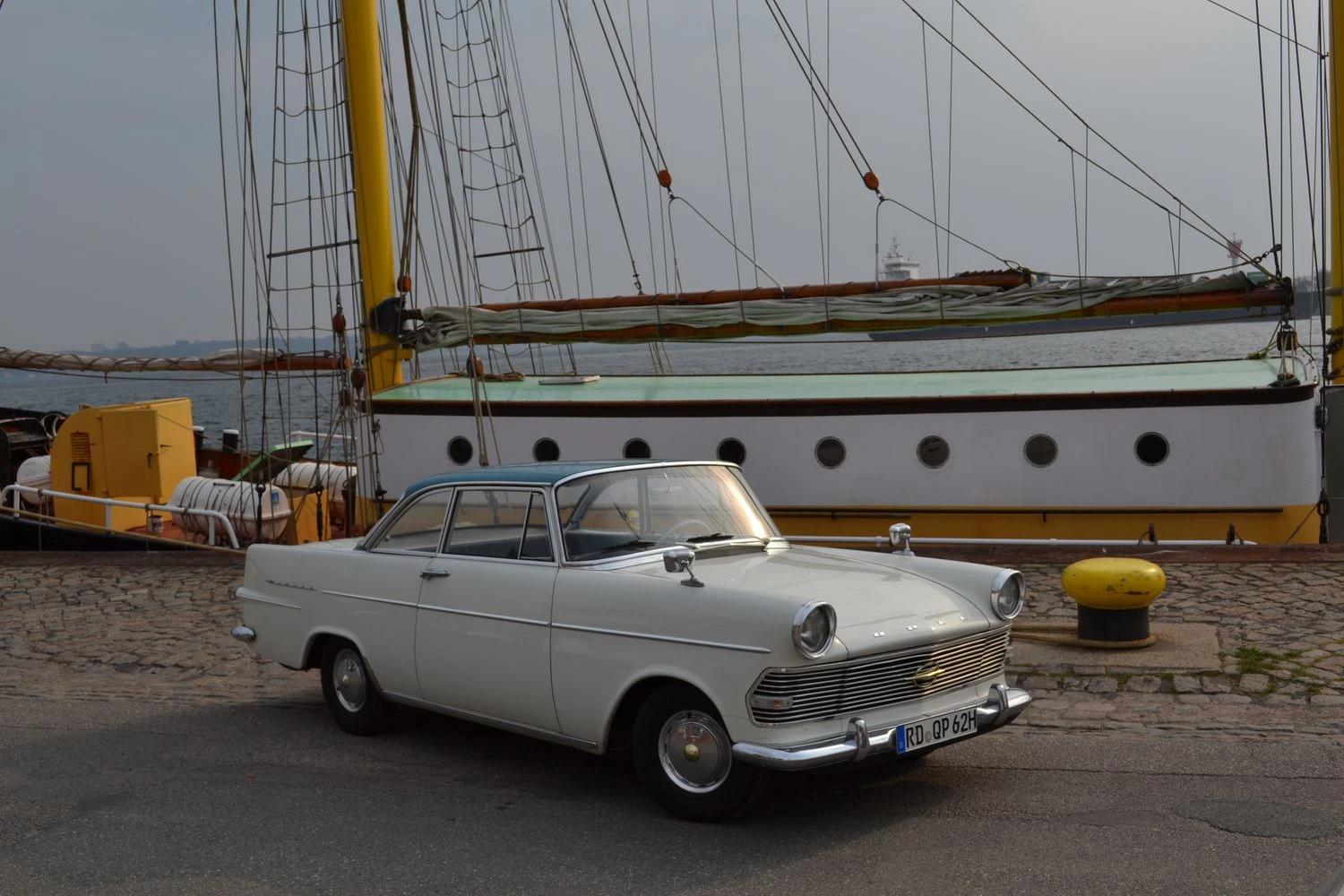 Ein weißes Oldtimer-Auto auf einer Kopfsteinpflasterstraße neben einem großen Segelschiff im Hafen.