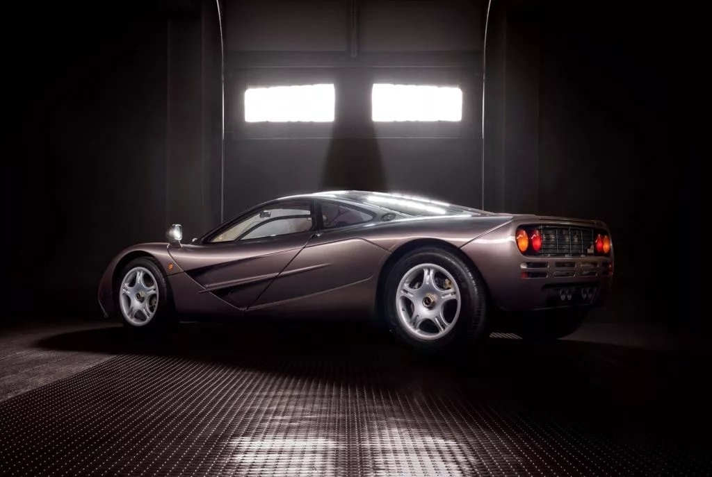 Ein brauner McLaren F1 steht in einer dunklen Garage. Das Auto ist seitlich von links nach rechts ausgerichtet und wird von oben beleuchtet, wodurch Reflexionen auf der Karosserie sichtbar sind.