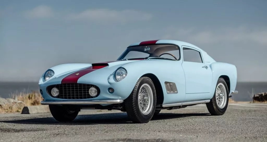 Hellblauer Oldtimer-Sportwagen mit rotem Mittelstreifen, runden Scheinwerfern und Speichenfelgen auf Asphalt vor bewölktem Himmel.