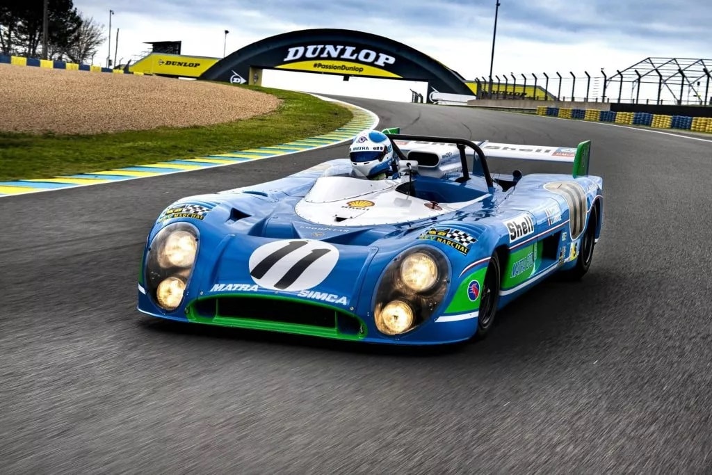 Blauer Rennwagen mit der Nummer 11 und 'MATRA-SIMCA'-Aufschrift auf der Motorhaube, fährt auf einer Rennstrecke mit Dunlop-Brücke im Hintergrund.