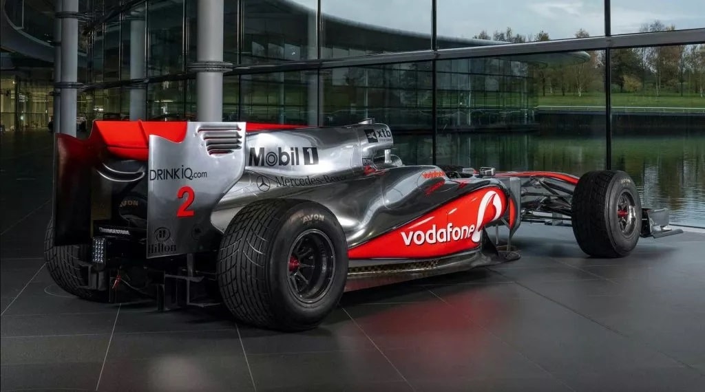 Formel-1-Rennwagen mit Vodafone- und Mobil-1-Branding, ausgestellt auf Fliesenboden in einem Raum mit großen Fenstern und Blick auf Bäume und Wasser.