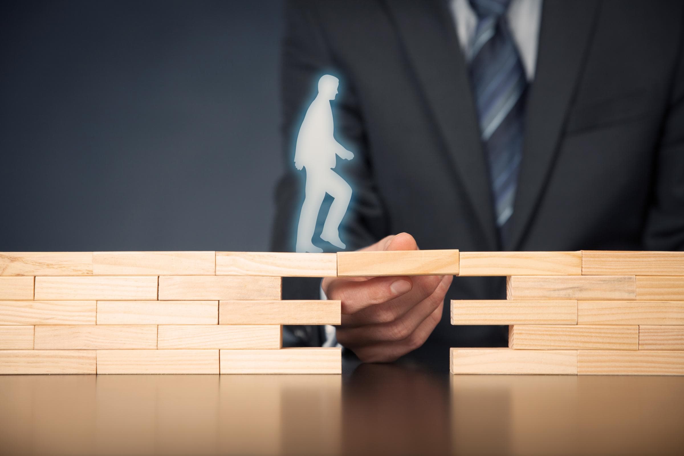 Person im Anzug legt einen Holzblock zwischen zwei Stapel, auf dem eine leuchtende Silhouette eine Treppe hinaufgeht – Symbol für Fortschritt.