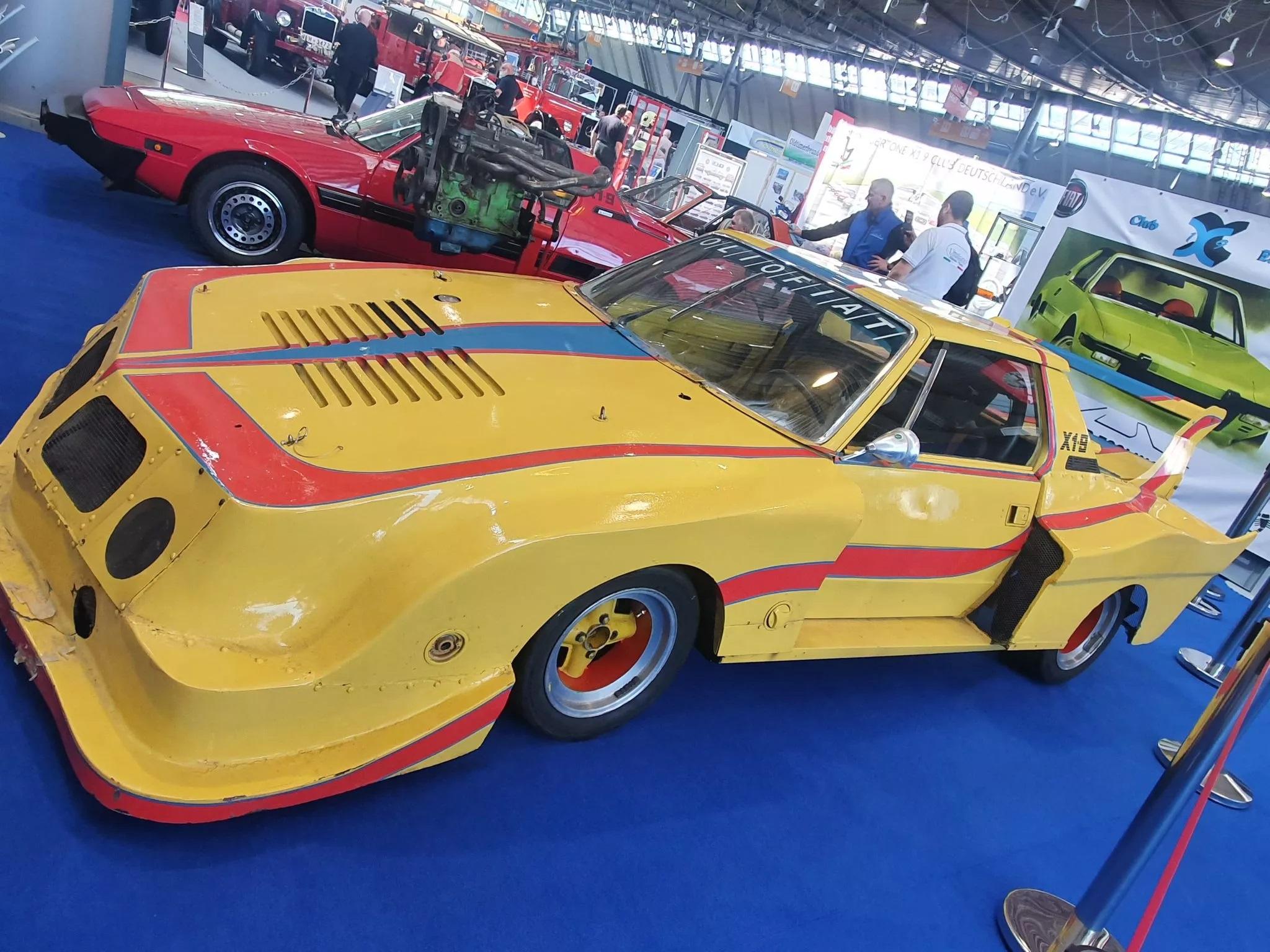 Gelber Rennwagen mit der Nummer X19 und roten Akzenten auf einer Messe, im Hintergrund ein rotes Auto mit offenem Motor.